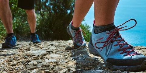 Nahaufnahme von zwei Personen, die auf einem steinigen Pfad wandern. Der Fokus liegt auf ihren Schuhen. Die Person im Vordergrund trägt graue Wanderschuhe mit roten Schnürsenkeln. Der Text auf dem Bild lautet „WANDERSCHUHE PFLEGEN“. Im Hintergrund sind blaues Wasser und grünes Laub zu sehen.