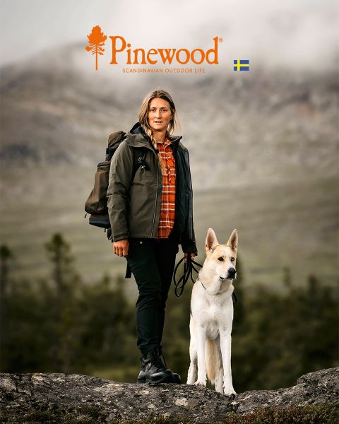 Ein Mann und eine Frau in Outdoor-Kleidung gehen unter einem bewölkten Himmel durch eine Wiese. Die Frau trägt eine schwarze Tasche, während der Mann einen Hut hält. Über ihnen sind das Pinewood-Logo und der Text „Scandinavian Outdoor Life“ zu sehen.