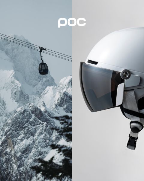 Eine Person in orangefarbenem Wintermantel und Helm steht auf einem schneebedeckten Berggipfel und blickt auf die fernen schneebedeckten Berge. Das POC-Logo ist auf dem Helm und in der oberen rechten Ecke des Bildes zu sehen.