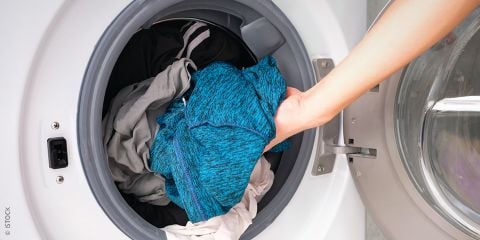Eine Hand legt bunte Sportkleidung, darunter ein blaues Shirt, in eine Frontlader-Waschmaschine. Über dem Bild ist der Text „Funktionskleidung pflegen“ zu sehen.