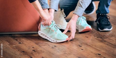 Eine Person in Jeans probiert einen hellgrünen Laufschuh mit weißer Sohle an. Eine andere Person hilft. Der Text lautet „Laufschuhe wie angegossen“ vor einem Holzbodenhintergrund.