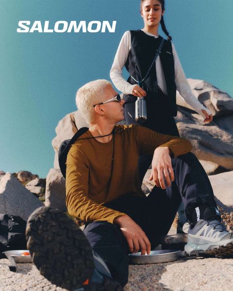 Nahaufnahme einer Person in Salomon-Schuhen und Salomon-Socken mit Farbverlauf in Blau. Sie steht auf einer Wiese mit einer malerischen Berglandschaft im Hintergrund. Ein Wanderer in Outdoor-Kleidung steht weiter entfernt, ebenfalls auf der Wiese.