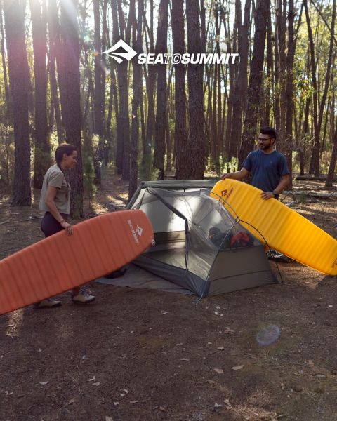 Eine Person liegt auf einer Isomatte, eingewickelt in einen gelben Schlafsack, und beobachtet einen Sonnenaufgang über einer Küstenklippe. Campingausrüstung liegt neben ihr, und die Meereswellen brechen gegen die Felsen. Das Logo „SEATOSUMMIT“ befindet sich in der unteren rechten Ecke.