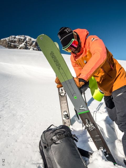 Ein Skifahrer in orangefarbener Jacke und Skibrille präpariert grüne Skier auf einem verschneiten Berghang, mit einem schwarzen Rucksack auf dem Boden und einem klaren blauen Himmel im Hintergrund.