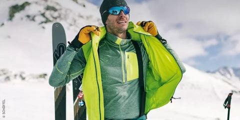 Eine Person in Wintersportkleidung steht in einer verschneiten Bergregion und hält eine hellgrüne Jacke offen, unter der sich eine graue Schicht mit Reißverschluss befindet. Sie trägt eine Mütze, eine Sonnenbrille und gelbe Handschuhe. Im Hintergrund sind Skier zu sehen. Der Text lautet „Schichten im Winter“.