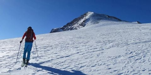 Eine Person in Winterkleidung fährt bei Sonnenschein einen schneebedeckten Berg hinauf. In der oberen linken Ecke ist der Text „SICHERHEIT AM BERG“ zu sehen.
