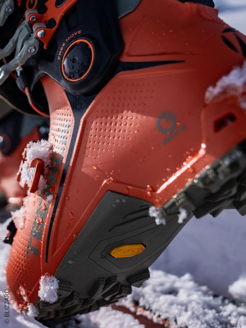 Nahaufnahme eines orangefarbenen Skischuhs, an dessen Sohle Schnee klebt, mit Details der strukturierten Oberfläche, der Schnallen und des Profils. Der Schuh steht auf einer verschneiten Fläche mit unscharfem Hintergrund.