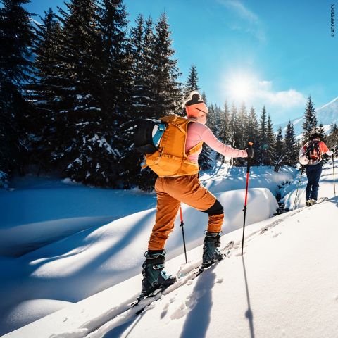 Eine Frau in Winterausrüstung und mit Rucksack fährt auf Skiern bergauf durch tiefen Schnee in einem sonnenbeschienenen, verschneiten Wald mit immergrünen Bäumen und einem strahlend blauen Himmel. Ein orangefarbenes Grafik-Overlay bedeckt die linke Seite des Bildes.