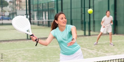 Frau spielt Padel-Tennis auf einem Außenplatz und bereitet sich darauf vor, einen Ball mit einem Schläger zu schlagen. Im Hintergrund ist eine weitere Spielerin zu sehen. Der Text lautet: „TRENDSPORT PADEL-TENNIS.“.