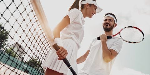 Ein Mann und eine Frau in weißer Sportkleidung stehen lächelnd auf einem sonnigen Tennisplatz. Beide halten Tennisschläger in den Händen. In der oberen linken Ecke ist der Text „FIT ZU ZWEIT“ zu sehen.