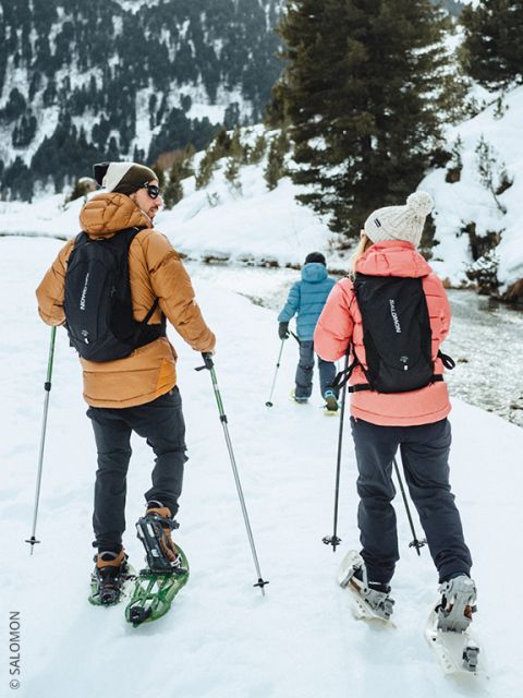 Drei Personen mit Winterjacken und Rucksäcken gehen mit Schneeschuhen auf einem verschneiten Weg entlang eines von Bäumen gesäumten Flusses, mit Bergen im Hintergrund.