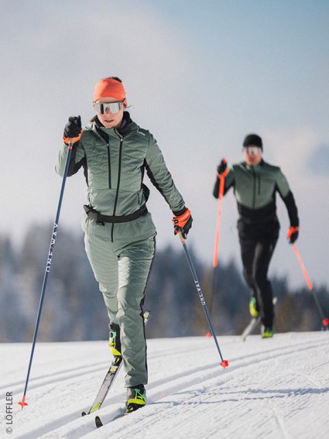Zwei Personen in grüner und schwarzer Kleidung mit orangefarbenen Akzenten beim Skilanglauf in einer verschneiten Landschaft. Die vordere Person trägt eine Sonnenbrille und eine Kapuze; im Hintergrund sind Bäume und ein blauer Himmel zu sehen.