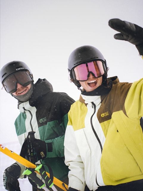 Zwei Personen in Skikleidung und Helmen lächeln und posieren auf einem verschneiten Berg. Einer hält gelbe Skier, während der andere in die Kamera winkt. Beide tragen grüne, weiße und gelbe Skijacken und Skibrillen.