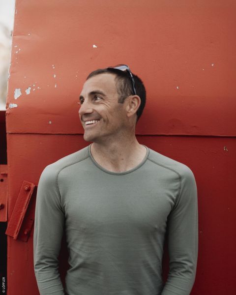 Ein lächelnder Mann in einem grünen Hemd steht vor einer großen roten Metallkonstruktion und schaut mit einer Sonnenbrille auf dem Kopf zur Seite.