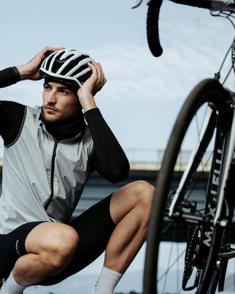 Ein Radfahrer mit Helm und Radfahrerkleidung kniet neben einem Fahrrad und stellt seinen Helm ein, mit einer Brücke und dem Himmel im Hintergrund.