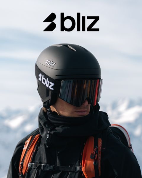 Eine Person, die einen schwarzen Skihelm und eine verspiegelte Skibrille trägt, beide mit der Aufschrift "Bliz" versehen, steht im Freien mit verschneiten Bergen im Hintergrund. Das Bliz-Logo ist am oberen Rand des Bildes gut sichtbar angebracht.