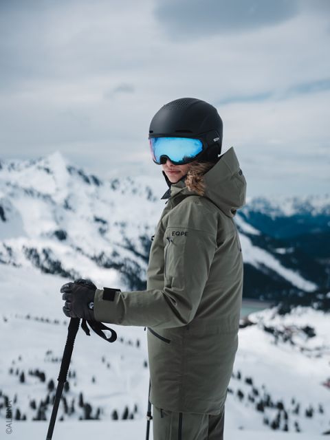 Ein Skifahrer in grüner Jacke und schwarzem Helm steht auf einem verschneiten Berghang, hält die Skistöcke und blickt zurück, mit Gipfeln im Hintergrund.