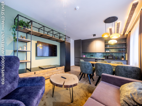 Moderner Wohnbereich mit lilafarbenem Sessel, lilafarbenem Sofa, rundem Couchtisch aus Holz, an der Wand montiertem Fernseher, offenen Regalen und einem Essbereich mit grünen Stühlen und Pendelleuchten neben einer eleganten Küche.