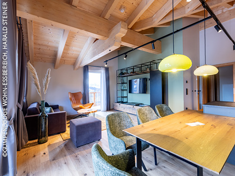 Ein moderner Wohn- und Essbereich mit einer Holzdecke mit sichtbaren Balken, großen Fenstern, gemütlichen Sitzgelegenheiten, einem an der Wand montierten Fernseher, einem Esstisch aus Holz und Pendelleuchten schafft eine warme und einladende Atmosphäre.