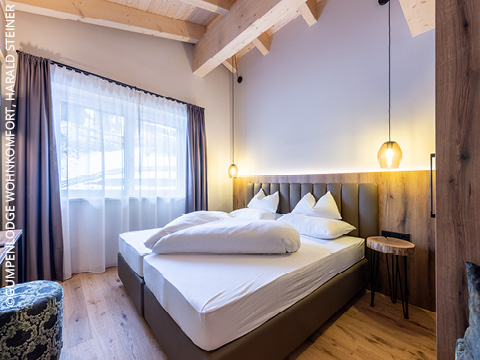 Modernes Schlafzimmer mit Doppelbett, weißem Bettzeug, Holzfußboden und sichtbaren Holzbalken. Ein großes Fenster mit transparenten und dunklen Vorhängen lässt natürliches Licht herein. Zwei Pendelleuchten hängen über dem Kopfteil.