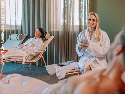 Drei Personen in weißen Gewändern entspannen sich auf Liegestühlen in einem Wellnessbereich. Eine Frau im Vordergrund lächelt und hält ein Getränk in der Hand, während sich eine andere im Hintergrund zurücklehnt. Sanfte Beleuchtung und Vorhänge schaffen eine ruhige Atmosphäre.