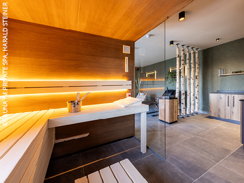 Moderner Saunaraum mit Holzbänken, einem Saunakübel, Handtüchern und sanfter Akzentbeleuchtung. Eine Glaswand trennt die Sauna von einem Entspannungsbereich mit Birkendekor und Holzschränken.