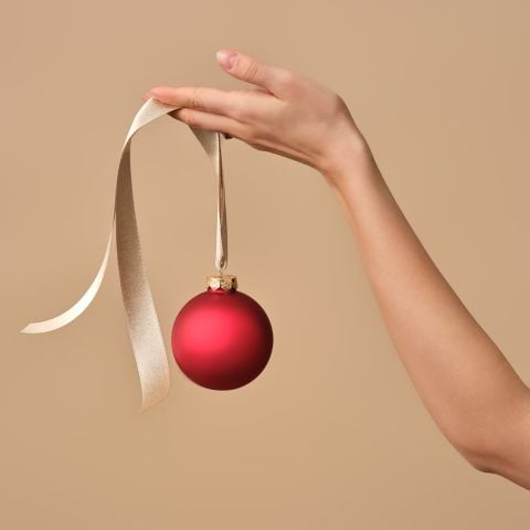 Eine Hand hält ein rotes Weihnachtsornament an einem beigen Band vor einem schlichten beigen Hintergrund.