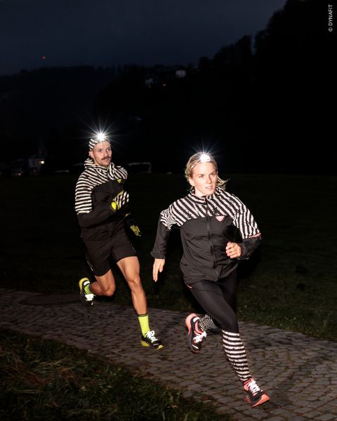 Zwei Personen laufen nachts mit Scheinwerfern und schwarz-weiß gestreiften Jacken im Scheinwerferlicht über einen Kopfsteinpflasterweg.