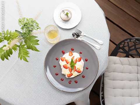 Ein Tisch mit einem Dessert mit Erdbeeren und Minze, einem Getränk, Löffel und Gabel, einem kleinen Kuchen und einer Vase mit grünen Blättern und Blumen.