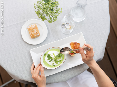 Eine Person hält einen Löffel über eine Schüssel mit grüner Suppe mit Rucola, serviert mit Brot und Gebäck auf einem weißen Tischtuch.