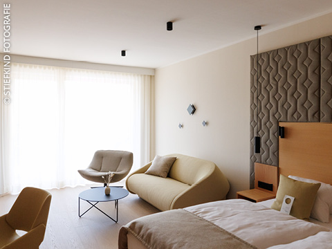 Modernes Hotelzimmer mit großem Bett, beigem Sofa, Sessel, rundem Couchtisch und bodentiefen Fenstern mit transparenten Vorhängen.