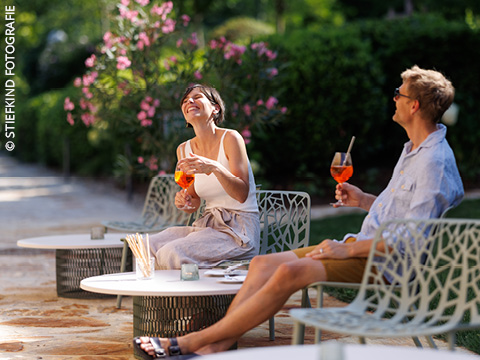 Ein Mann und eine Frau sitzen draußen auf Gartenstühlen, lächeln und halten orangefarbene Getränke (Aperol) in der Hand, umgeben von Grün und rosa Blumen an einem sonnigen Tag.