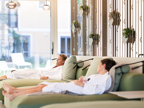 Zwei Personen in weißen Bademänteln entspannen sich auf Loungesesseln in einem Raum mit Topfpflanzen an der Wand und großen Fenstern, die Tageslicht hereinlassen.