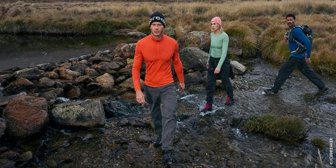 Drei Personen in Outdoor-Ausrüstung überqueren einen felsigen Bach in einer grasbewachsenen, bergigen Gegend, wobei eine Person vorausgeht und die anderen hinterhergehen.