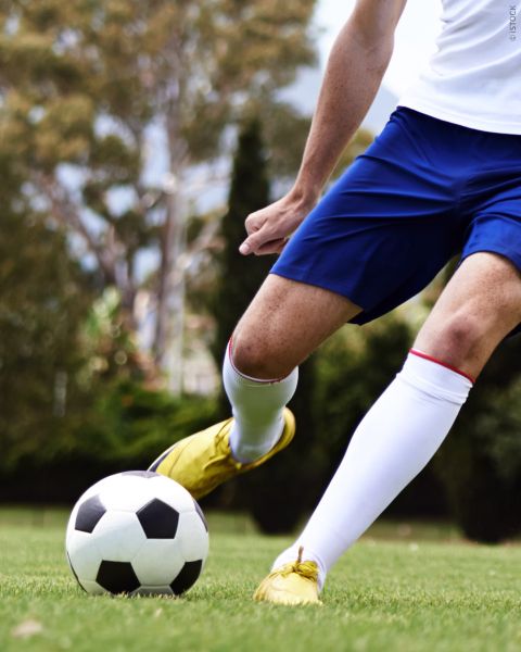 Fußballspieler in blauen Shorts und gelben Schuhen, der einen schwarz-weißen Fußball auf einer Wiese kickt, mit Bäumen im Hintergrund.