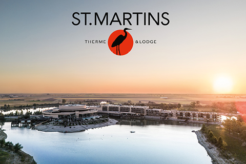 Luftaufnahme der St. Martins Therme & Lodge an einem See bei Sonnenuntergang, mit dem Logo und Namen des Resorts über der Szene.