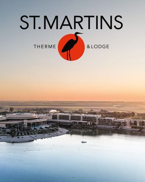 Luftaufnahme des Resorts St. Martins Therme & Lodge an einem See bei Sonnenuntergang, mit modernen Gebäuden und einem Vogellogo darüber.