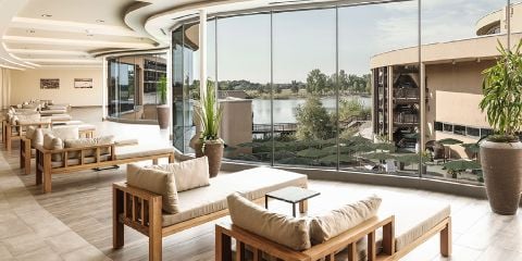 Modernes Wohnzimmer mit Holzmöbeln und beigen Kissen, große Fenster mit Blick auf den Fluss und die Außenanlagen, mit Topfpflanzen im Zimmer.