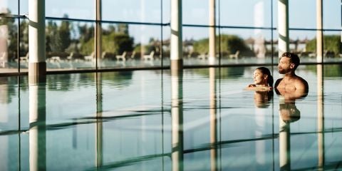 Ein Mann und eine Frau entspannen sich gemeinsam in einem Hallenbad mit großen Fenstern und einer schönen Aussicht auf die Umgebung.