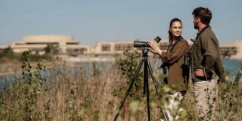 Zwei Personen, die mit Spektiv und Fernglas an einem See Vögel beobachten, mit Gebäuden im Hintergrund und hohem Gras im Vordergrund.