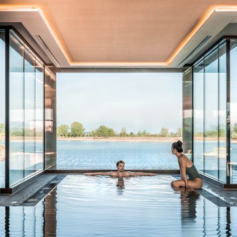 Zwei Personen entspannen sich an einem Innenpool mit Glaswänden, der einen Blick auf einen ruhigen See und Bäume unter einem klaren Himmel bietet.