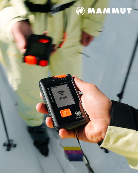 Nahaufnahme einer Person, die ein Mammut-Lawinenverschüttetensuchgerät hält, das den Modus "SEND" anzeigt, mit einer anderen Person und verschneitem Boden im Hintergrund.