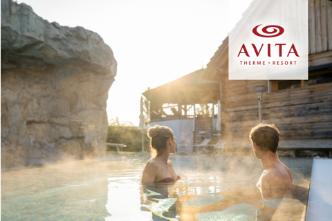 Ein Mann und eine Frau entspannen sich in einem dampfenden Außenpool eines Resorts, mit dem Logo des AVITA Therme Resorts in der oberen rechten Ecke.