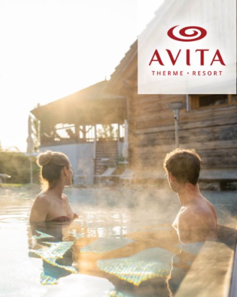 Ein Mann und eine Frau entspannen sich in einem dampfenden Außenpool im Avita Therme Resort, mit einem rustikalen Holzgebäude im Hintergrund.