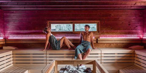 Zwei Personen entspannen sich in einer Holzsauna, auf Bänken sitzend und mit Handtüchern umwickelt; im Vordergrund sind Felsen zu sehen, im Hintergrund ist ein Fenster zu sehen.