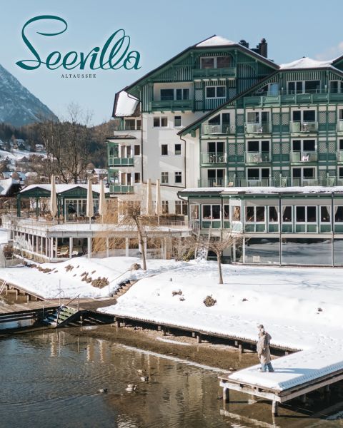 Hotel am See mit grünen Balkonen und schneebedeckter Umgebung; eine Person steht auf einem Steg, mit Bergen im Hintergrund.