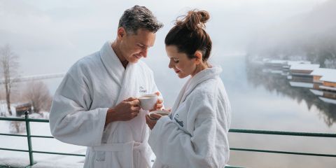 Ein Mann und eine Frau in weißen Gewändern genießen heiße Getränke auf einem Balkon mit Blick auf einen verschneiten, ruhigen See und Holzhütten.