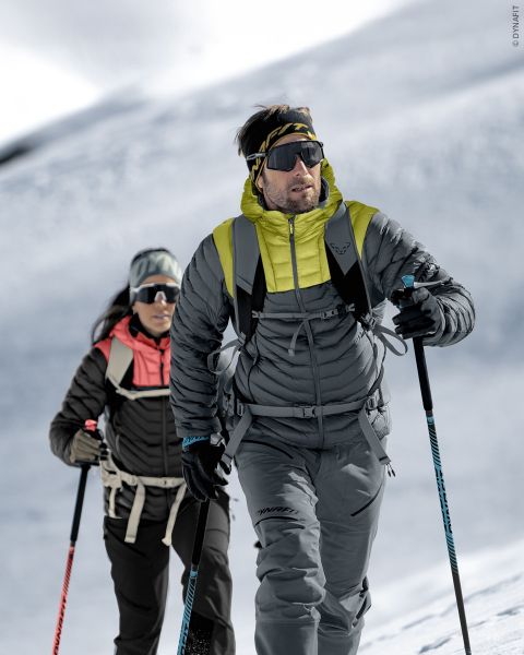 Zwei Personen in Winterausrüstung wandern mit Skistöcken, Sonnenbrillen und Rucksäcken unter einem hellen, bewölkten Himmel einen verschneiten Berghang hinauf.