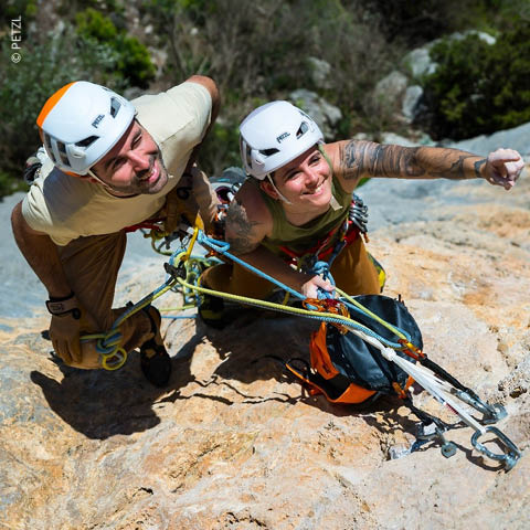 Zwei Kletterer mit Helmen überprüfen Kletterausrüstung und Seile, während sie sich an einer Felswand festhalten und sich auf eine Klettertour bei sonnigem Wetter vorbereiten.