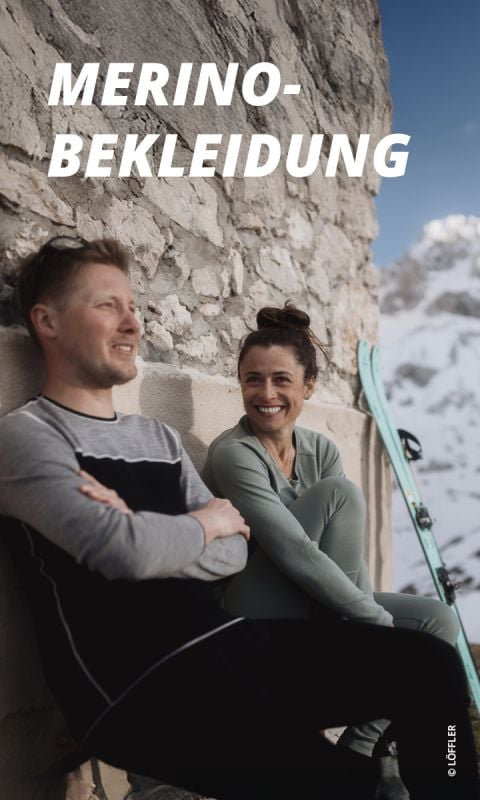 Ein lächelnder Mann und eine lächelnde Frau in Merinokleidung sitzen an einer Steinmauer mit Skiern, im Hintergrund verschneite Berge. Text: "MERINO-BEKLEIDUNG".
